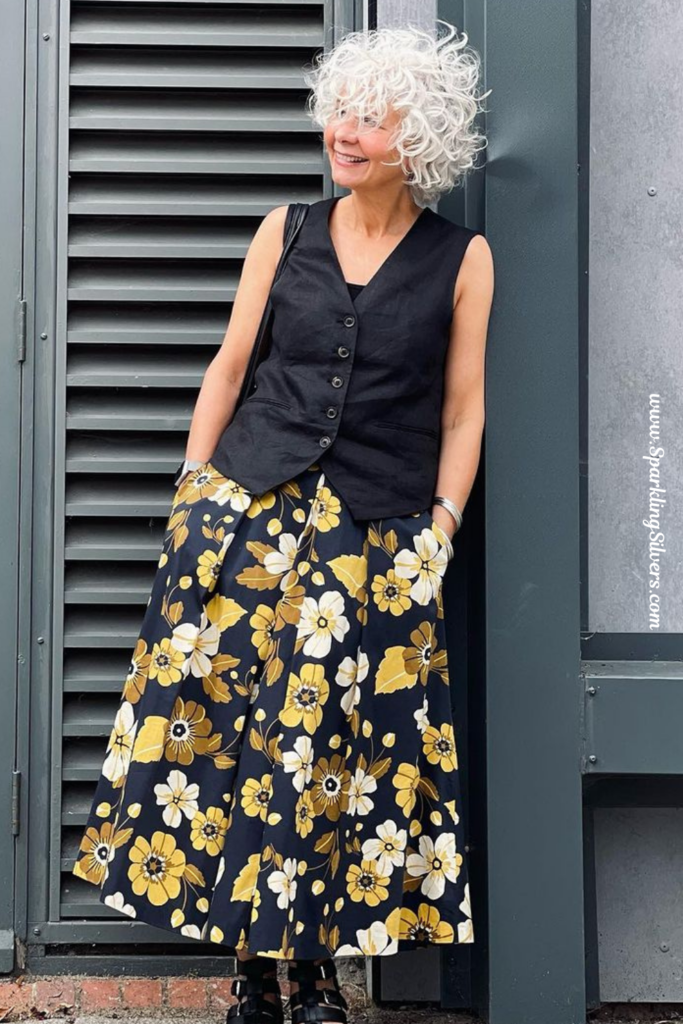 A Woman with white hair wearing a black waistcoat and a floral skirt.